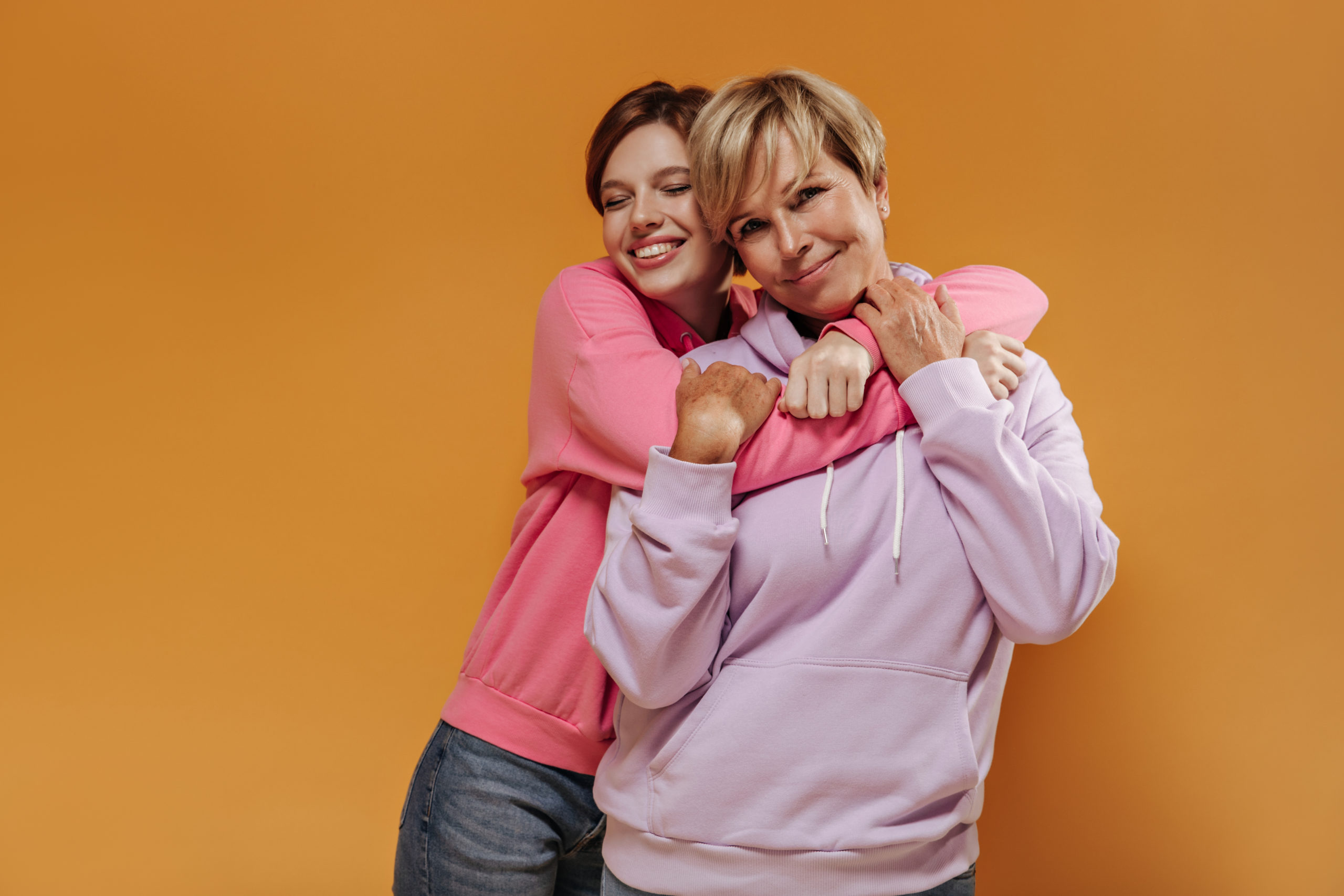 Lovely short haired girl in trendy hoodie and jeans smiling with closed eyes and hugging her mother in stylish clothes on orange backdrop.